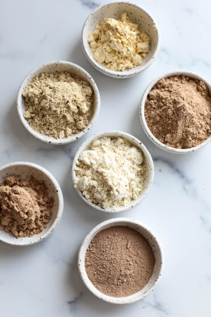 6 shallow white bowls filled with different protein powders. 