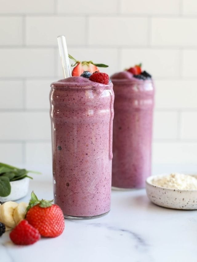 Two tall glasses of berry protein smoothies with fresh berries and protein powder in the forefront. 