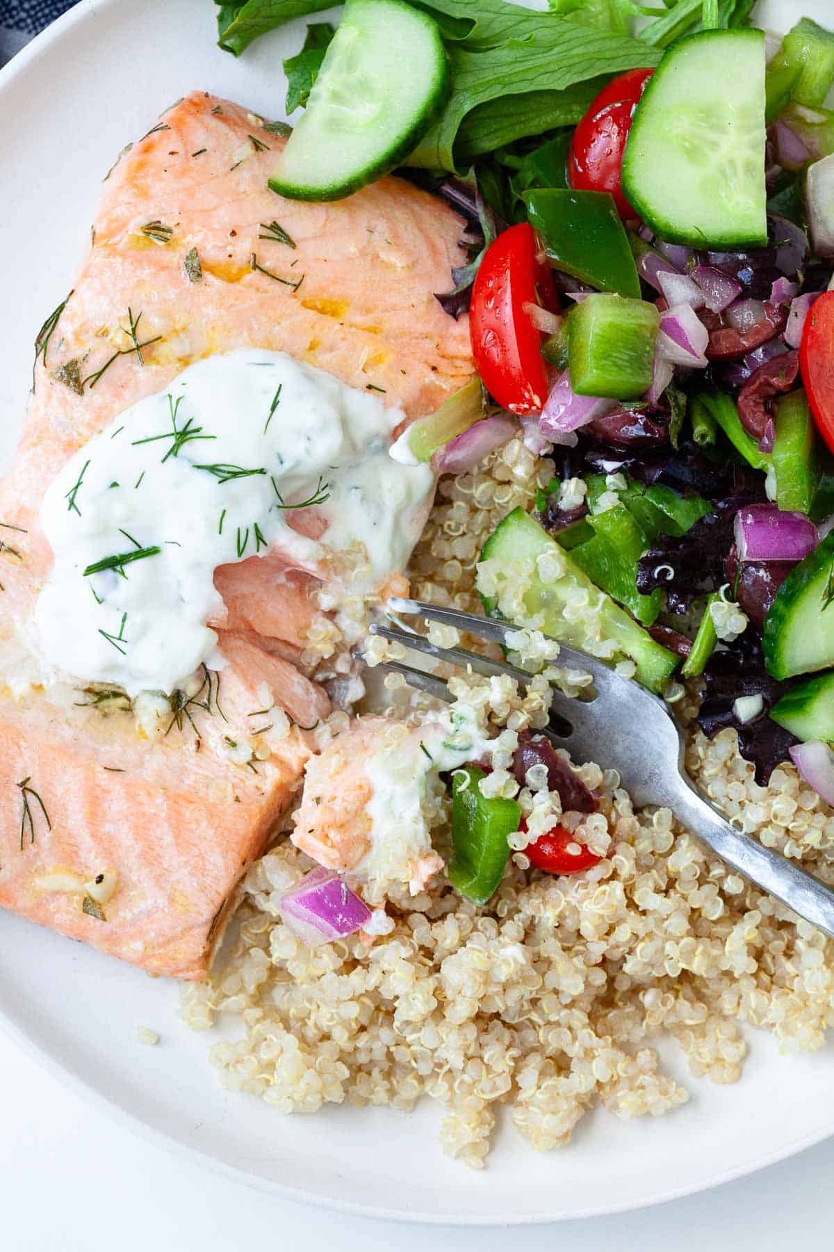 Flaked salmon in Greek salmon quinoa bowl with dollop of tzatziki, quinoa, and chopped vegetables.