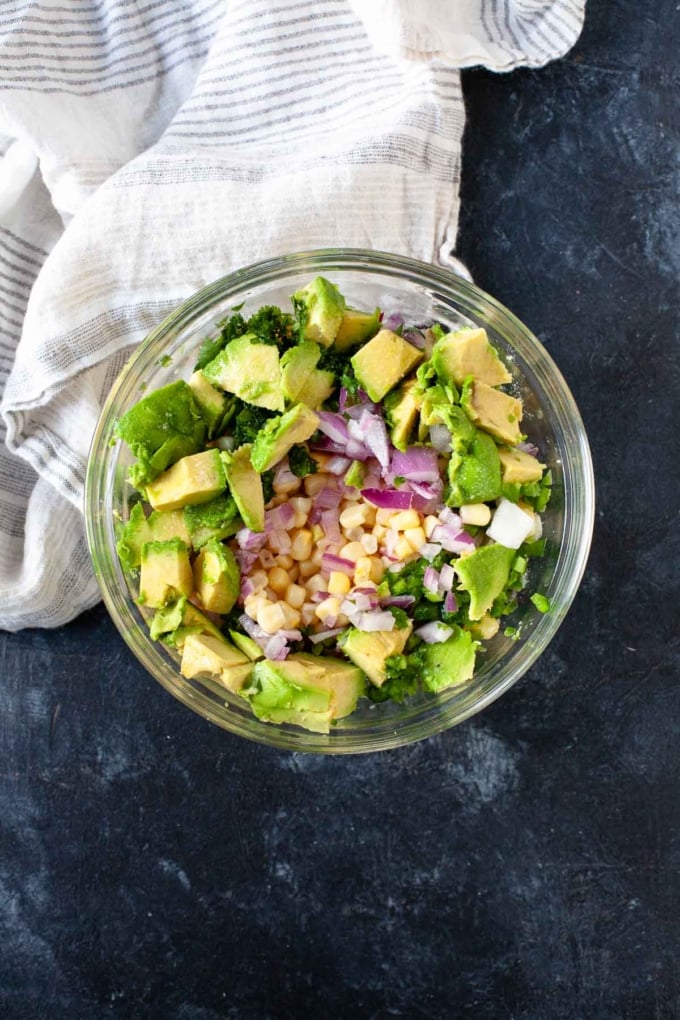 avocado, corn, red onion, cilantro in glass bowl on dark tabletop next to white striped dishtowel 