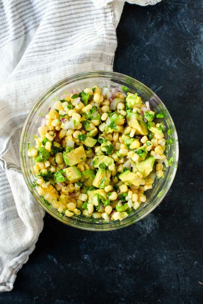 corn avocado salsa mixed together in glass bowl on dark tabletop next to white striped dish towel
