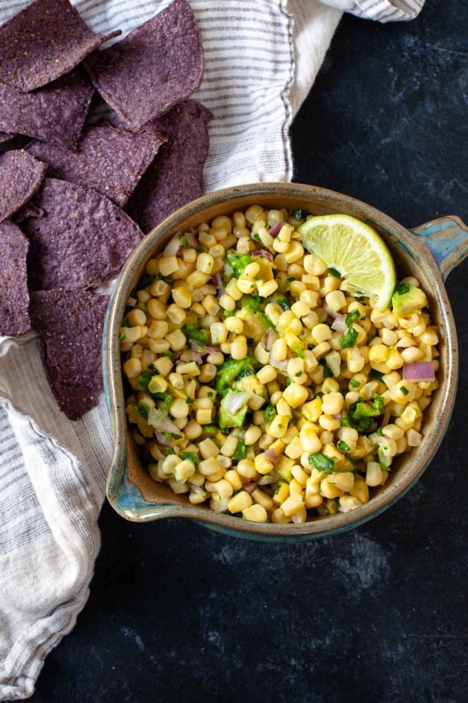 corn avocado salsa with lime wedge in brown and blue ceramic bowl next to blue tortilla chips on dish towel